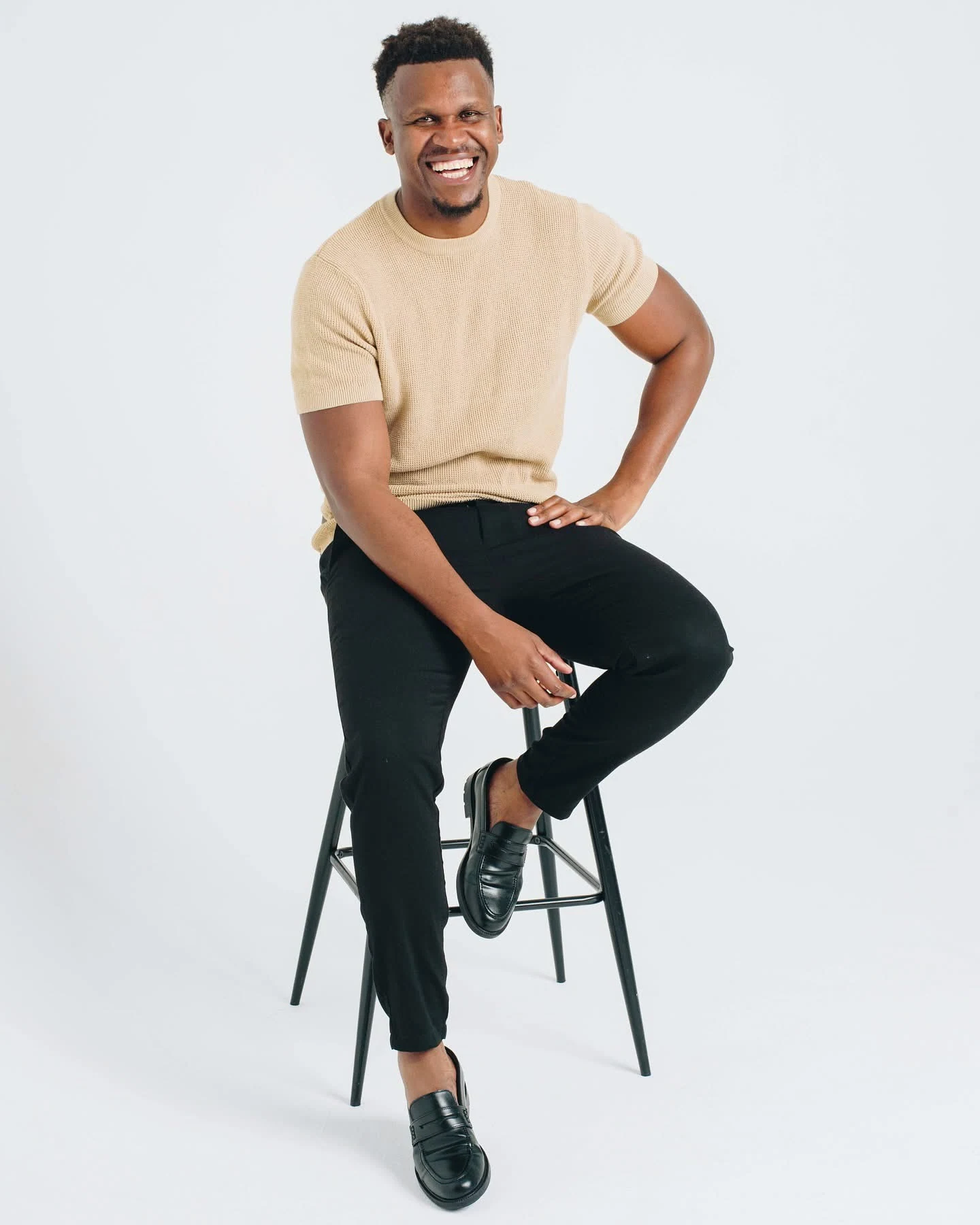 Phinius smiling, seated on a stool in a beige shirt.