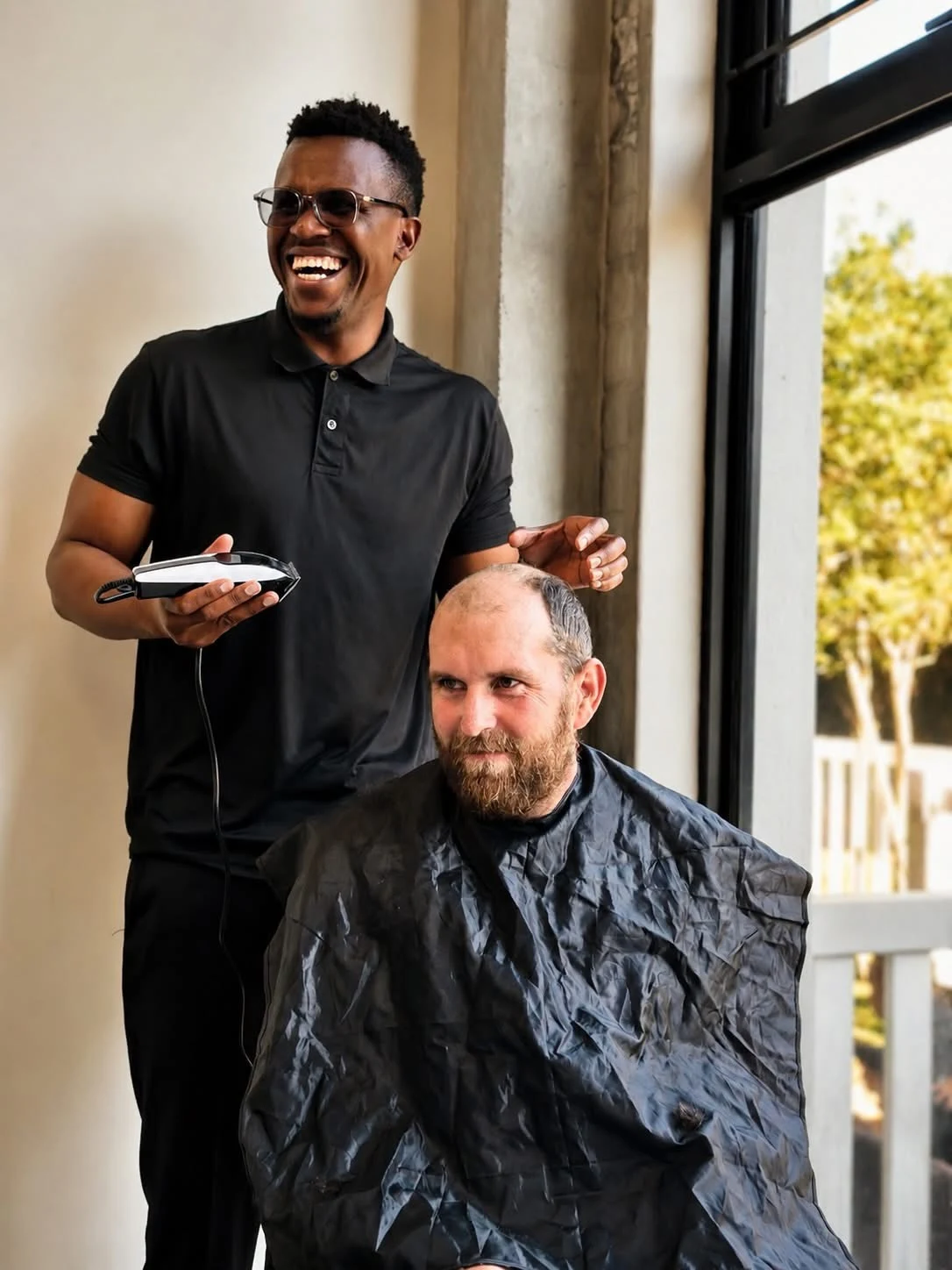 Phinius sharing a light-hearted moment while cutting a seated man’s hair.
